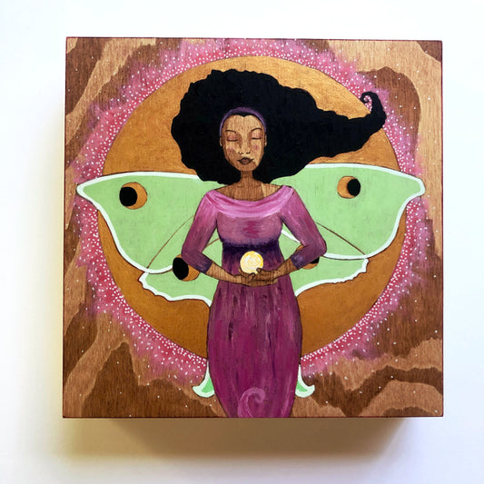 Art depicting a Black woman in a purple gown holding a ball of light.  She has the wings of a Luna moth on her back.  She is standing in front of a golden full moon.  The wood grain in the background is prominent.  
