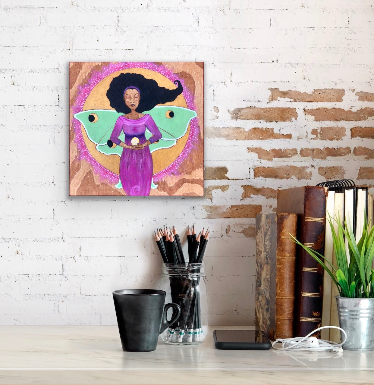 Art print hanging on a brick wall above books, pencils and other items on a desk.  The print by Portland artist Lea K. Tawd portrays a Black woman with wings of a Luna moth and a full moon behind her. 