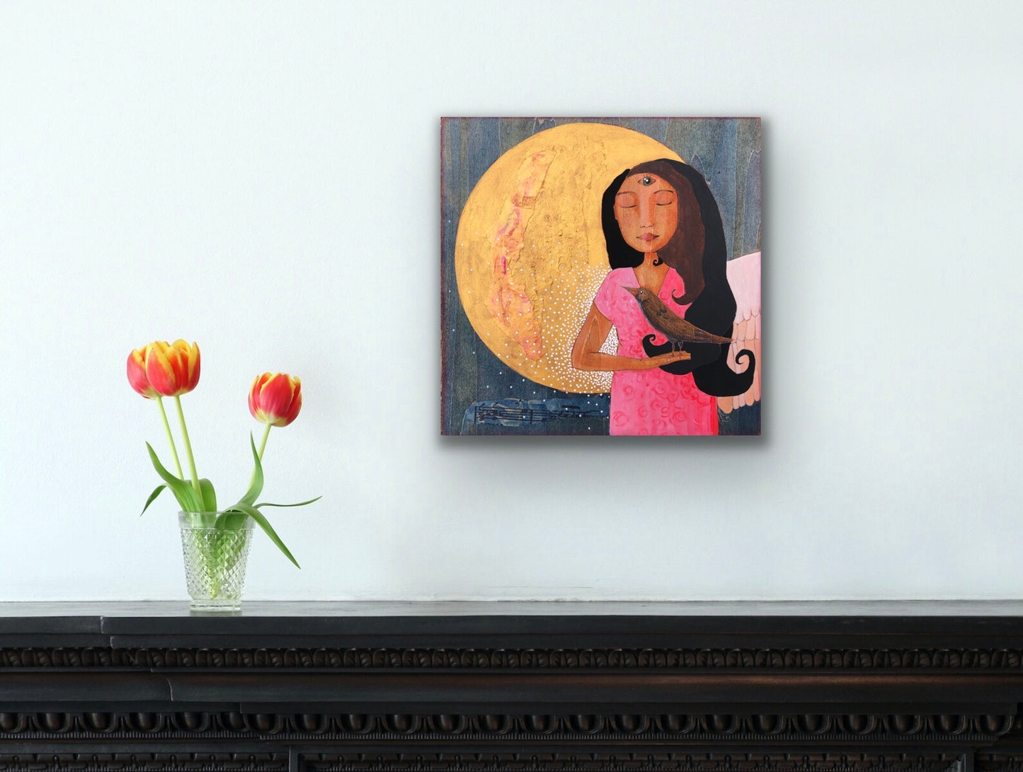 A small painting of a winged woman holding a magpie standing in front of a full moon with her 3rd eye open.  The painting hangs on a white wall above a dark shelf with a vase of tulips sitting on it. 