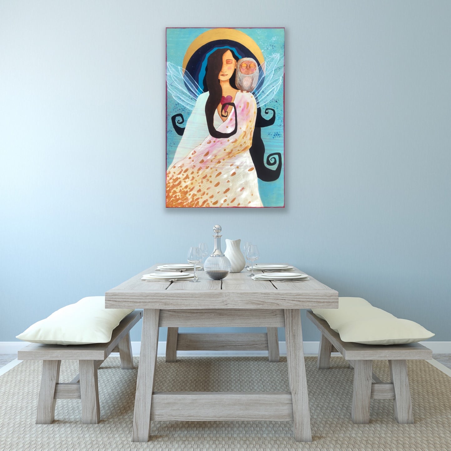 Beautiful spiritual artwork of a white and gold-robed woman with wings  and an owl perched on her shoulder.  The artwork is hanging on a light grey wall over a wooden dinner table with wooden benches and neutral settings and pillows.
