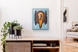small spiritual artwork of a woman and feathers on a blue background.  The painting is hanging on a white wall over a shelf with plants on it.