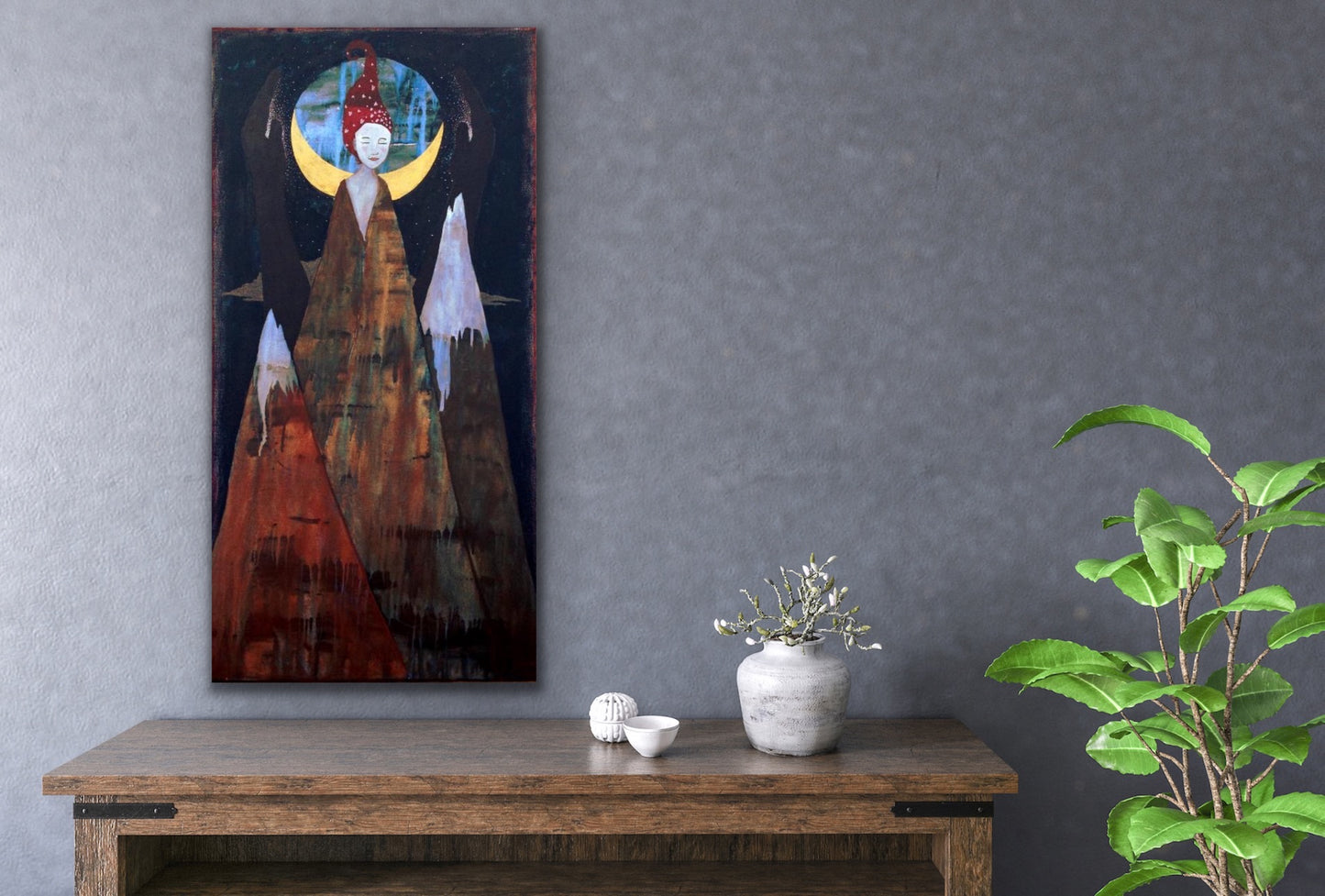 cosmic artwork depicting a woman formed from the top of a mountain. the painting is hanging on a gray wall above a wooden table with a plant and 3 white objects