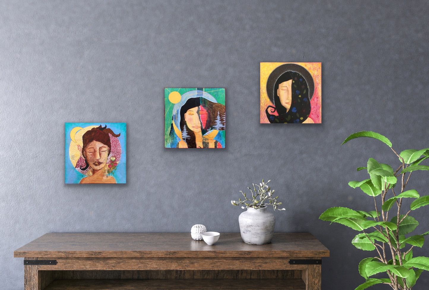 three vibrant, colorful mixed media feminine artworks hanging on a gray wall above a wooden table with white pots and a flower. beside the table is a large green plant